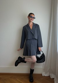 Conjunto cinza ajustado de duas peças com um blazer curto e uma saia plissada. Meias-calças de rede transparentes, botas pretas de tornozelo e uma bolsa preta em tecido trançado.