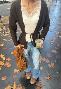 Osoba stoji na mokrom pločniku prekrivenom jesenskim lišćem, nosi smeđi kardigan, bijelu čipkastu bluzu, plave traperice i smeđe cipele, drži smeđu torbu i zeleni napitak.