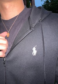 Melna kapuce ar rāvējslēdzēju, kurai kreisajā krūtī ir balta izšūta polo spēlētāja logo, uzvilkta virs tumša apaļā kakla krekla.