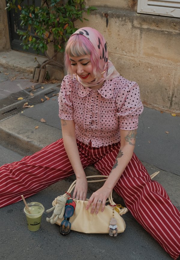 Joven mujer con cabello rosa y pañuelo en la cabeza sentada con las piernas cruzadas en la acera, lleva blusa de lunares y pantalones a rayas, sosteniendo una bolsa con llaveros.
