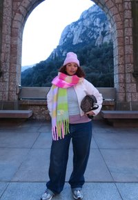 Femme portant un bonnet rose et une écharpe colorée, debout sous une arche en pierre avec un paysage montagneux en arrière-plan, tenant un sac à motifs.