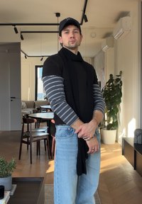 Άνδρας με μαύρο καπέλο, στρωμένη μαύρη και ριγέ μπλούζα, ανοιχτό μπλε τζιν και δαχτυλίδια στέκεται σε έναν φωτεινό μοντέρνο χώρο καθιστικού και τραπεζαρίας.