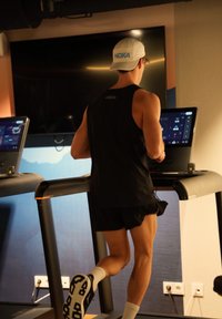 Une personne en débardeur noir et short court court sur un tapis de course doté d'un affichage numérique. Une casquette blanche HOKA orne sa tête.