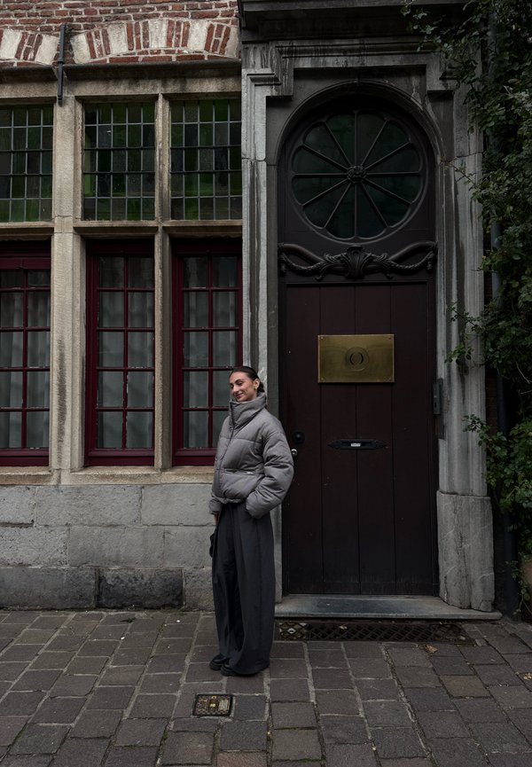 Une personne vêtue d’une veste grise et d’un pantalon foncé se tient à côté d’une entrée voûtée ornée sur un bâtiment historique en briques et en pierre.