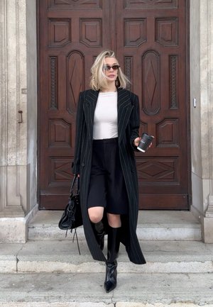 Mujer rubia con gafas de sol, abrigo negro a rayas, camiseta blanca, pantalones cortos negros y botas baja por escalones de piedra sosteniendo café y bolso.