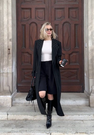 Mujer rubia con gafas de sol, abrigo negro a rayas, camiseta blanca, pantalones cortos negros y botas baja por escalones de piedra sosteniendo café y bolso.