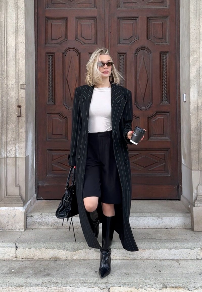 Femme blonde portant des lunettes de soleil, un manteau noir à rayures fines, un haut blanc, un short noir et des bottes descend des marches en pierre en tenant un café et un sac à main.
