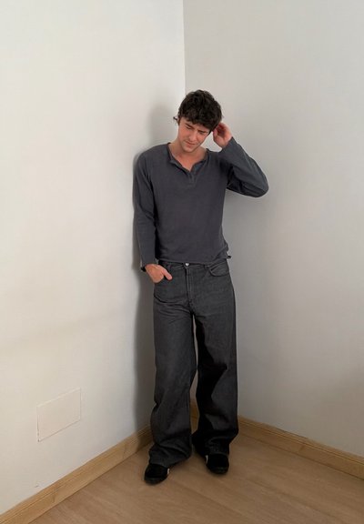 Joven con cabello rizado y oscuro, de pie contra una pared en esquina, vestido con una camisa oscura de manga larga y pantalones anchos negros, con una mano en el bolsillo.