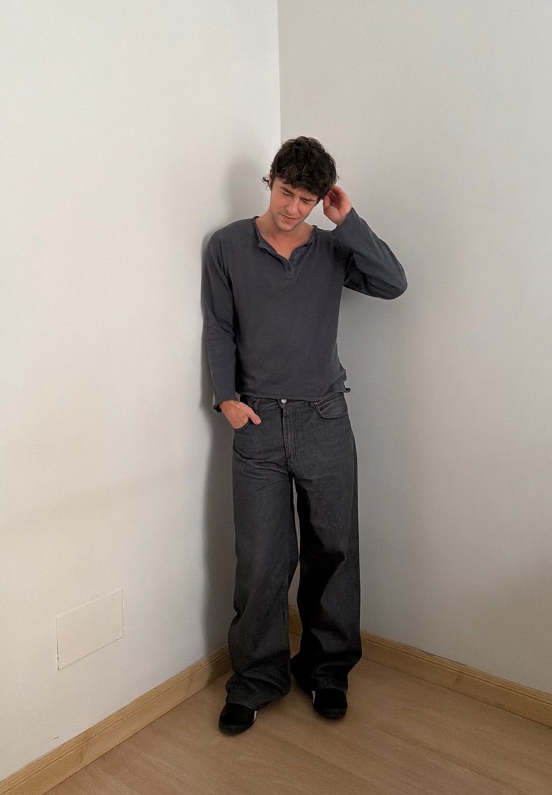 Jeune homme aux cheveux bouclés foncés se tenant contre un mur d'angle, portant une chemise à manches longues sombre et un large pantalon noir, une main dans la poche.