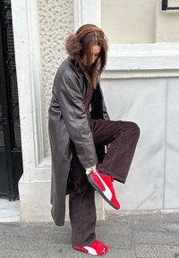 Femme portant un pantalon marron et un manteau gris, ajustant des baskets rouges et blanches tout en se tenant sur un trottoir en ville près d'un mur de couleur claire.