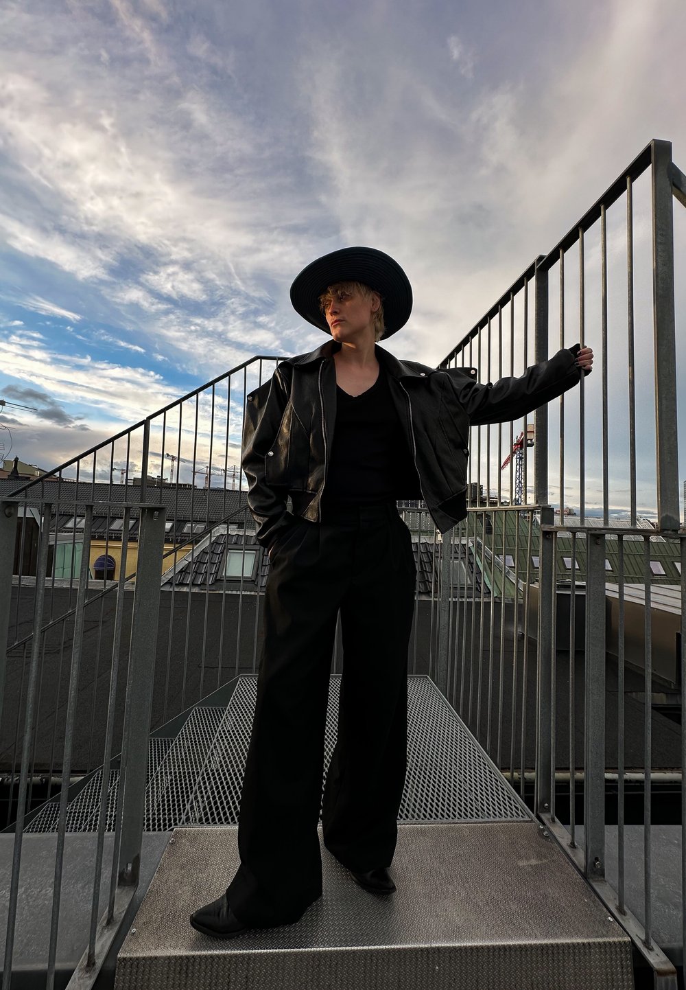 Μαύρο δερμάτινο μπουφάν με καπέλο με φαρδύ γείσο, μαύρο καρέ V και φαρδιά μαύρα παντελόνια. Μεταλλικό κράσπεδο και συννεφιασμένος ουρανός στο φόντο.