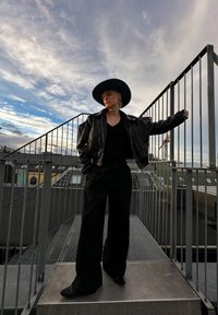 Μαύρο δερμάτινο μπουφάν με καπέλο με φαρδύ γείσο, μαύρο καρέ V και φαρδιά μαύρα παντελόνια. Μεταλλικό κράσπεδο και συννεφιασμένος ουρανός στο φόντο.