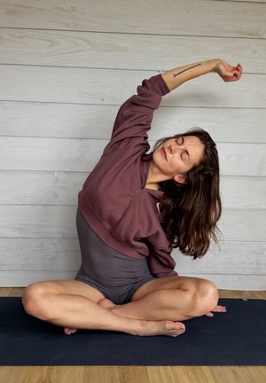 Mujer con sudadera granate y pantalones cortos grises sentada con las piernas cruzadas sobre una esterilla de yoga negra, estirando su brazo izquierdo por encima de la cabeza con los ojos cerrados.