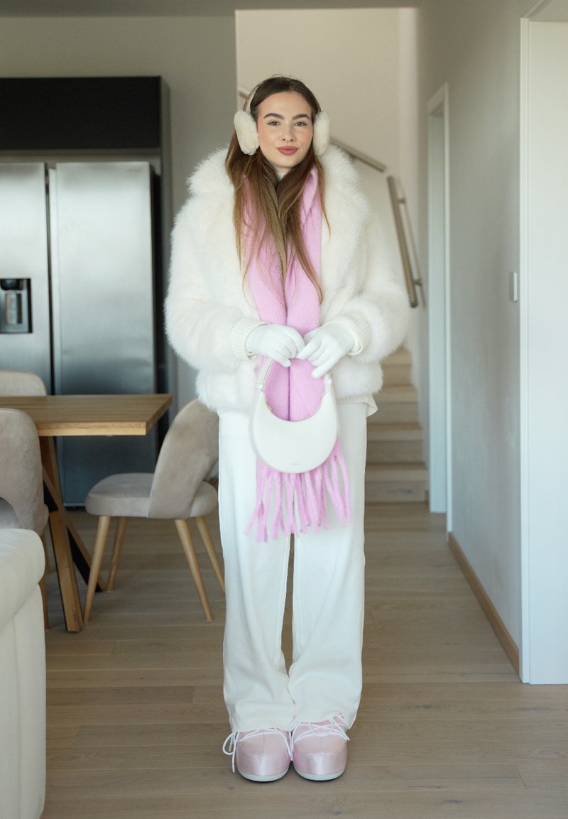 Jeune femme à l'intérieur portant un manteau en fourrure blanche, une écharpe rose, des cache-oreilles, des gants et des bottes, debout dans une cuisine et une salle à manger modernes.