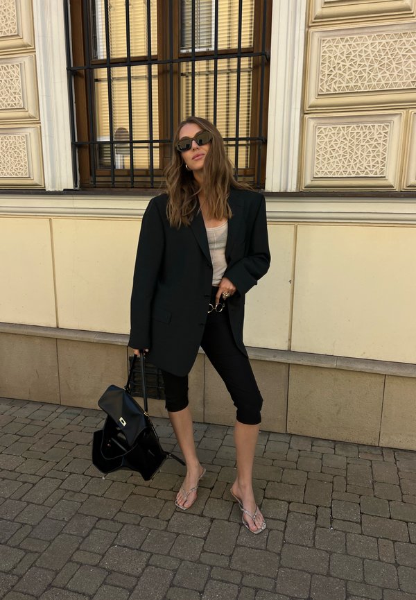 Žena v nadrozmernom čiernom saku, béžovom topu, čiernych skrátených nohaviciach a sandáloch stojí na dláždenej ulici a drží čiernu kabelku.