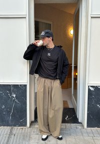 Sort lynlåstrøje, sort t-shirt med logo, beige bukser med vide ben og sorte og hvide sko. Holder en hvid kaffekop.