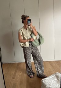 Beige κοντό γιλέκο με φερμουάρ, συνδυασμένο με φαρδιά παντελόνια με λιονταρίσιο σχέδιο. Μια πράσινη πλεκτή τσάντα κρατιέται στο ένα χέρι. Ο ξύλινος πάτωμα είναι ορατός.