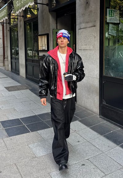 Joven caminando por la calle de la ciudad con una chaqueta de cuero negra, sudadera con capucha roja, pantalones negros sueltos, gorro multicolor y sosteniendo una taza de café en la mano derecha.