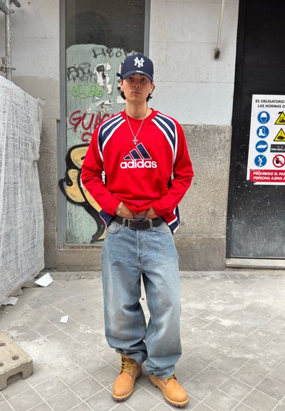 Joven con una camiseta roja de Adidas, vaqueros holgados, botas amarillas y una gorra azul marino de pie en una acera de la ciudad con grafitis y señales de seguridad detrás de él.