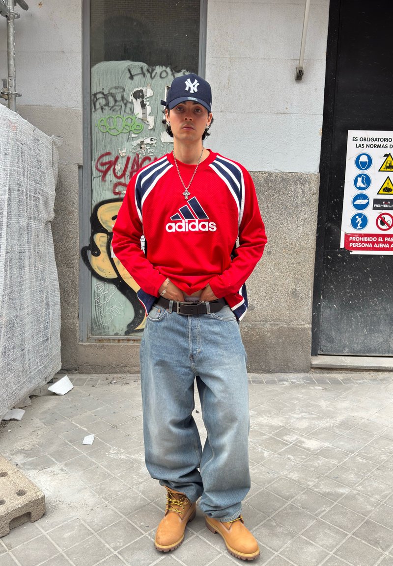 Jeune homme portant un t-shirt rouge Adidas, un jean ample, des bottes jaunes et une casquette bleu marine, debout sur un trottoir en ville avec des graffitis et des panneaux de sécurité derrière lui.