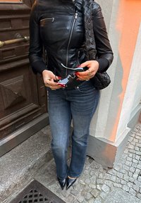 Fekete bőrkabát cipzárokkal, kék farmernadrág és fekete, hegyes orrú magassarkú cipő. Kezek fekete, ívelt szárú szemüveget tartanak. Városi környezet.