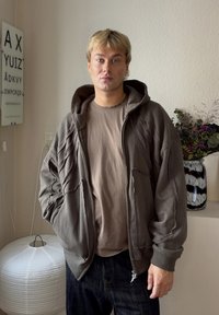 Sweat à capuche zippé gris foncé avec des détails texturés sur les manches, porté sur un t-shirt marron clair ; associé à un jean foncé. Lanterne en papier blanche en arrière-plan.