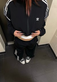 Casaco preto da Adidas com fecho, com riscas brancas, combinado com calças largas pretas e ténis chunky preto e branco.