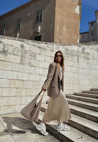 Blazer beige sobre uma blusa branca e saia maxi de seda bege, combinando com sapatilhas cinzentas. Transportando uma bolsa cinza, a descer escadas de pedra.