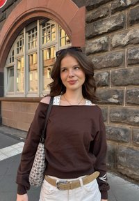 Brun genser med avskåret design, hvit prikkete cami under, beige belte og mønstret håndveske foran en steinvegg.