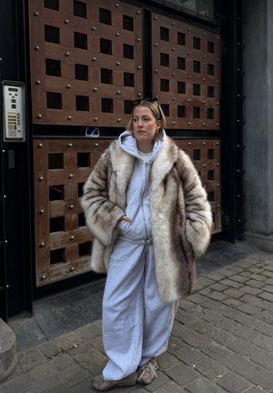 Mujer con un gran abrigo de piel sobre una sudadera gris con capucha y pantalones deportivos, de pie con las manos en los bolsillos en una calle empedrada frente a una puerta de madera.