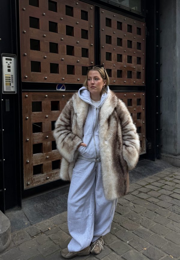 Donna che indossa un grande cappotto di pelliccia sopra una felpa grigia con cappuccio e pantaloni della tuta, in piedi con le mani nelle tasche su una strada di ciottoli davanti a un cancello di legno.