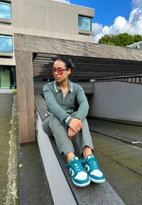 Ribbet grønn zip-up genser med hvit krage, grå bukser og teal-og-hvite joggesko, sittende på en grå kant mot en moderne bygning.