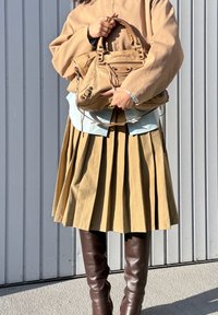 Sac à main en cuir beige avec poignée tissée, chemise bleu clair, jupe plissée beige et bottes marron montant au-dessus du genou sur un fond gris.