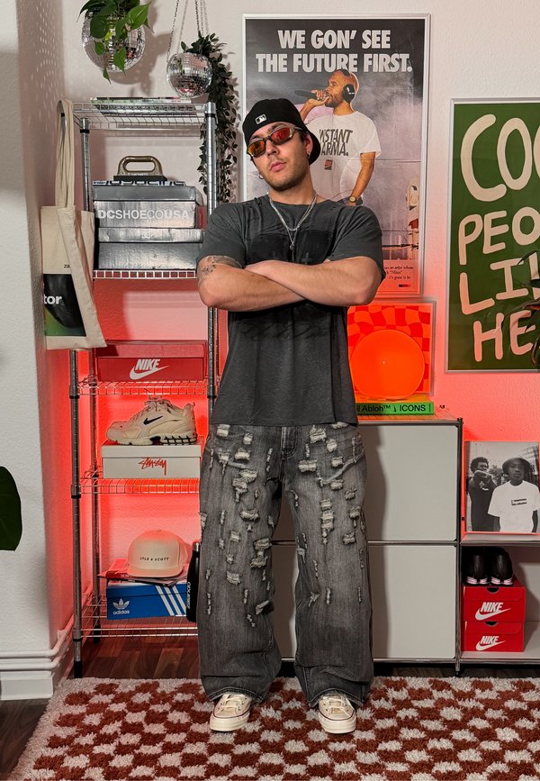 Muškarac sa prekriženim rukama, nosi poderane traperice, sunčane naočale i kapu, stoji u sobi s kutijama za tenisice i uokvirenim plakatima na zidovima.