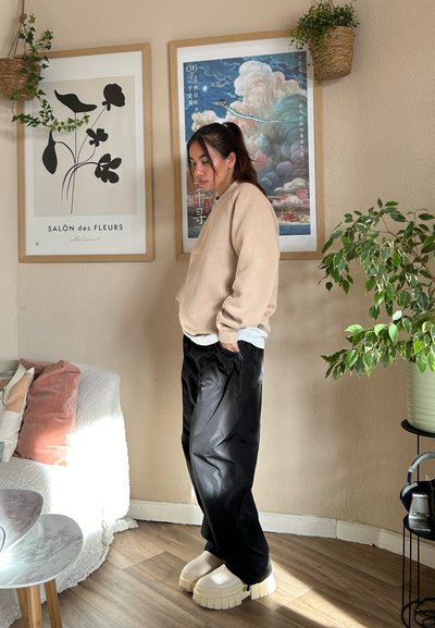 Sudadera beige, pantalones cargo negros con bolsillo con cremallera y zapatos de plataforma gruesa color crema. El fondo incluye arte enmarcado y vegetación.