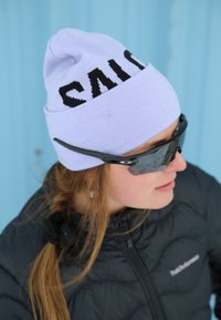 Personne portant des lunettes de soleil noires, un bonnet violet clair avec un texte noir, et une veste matelassée noire sur un fond bleu clair.