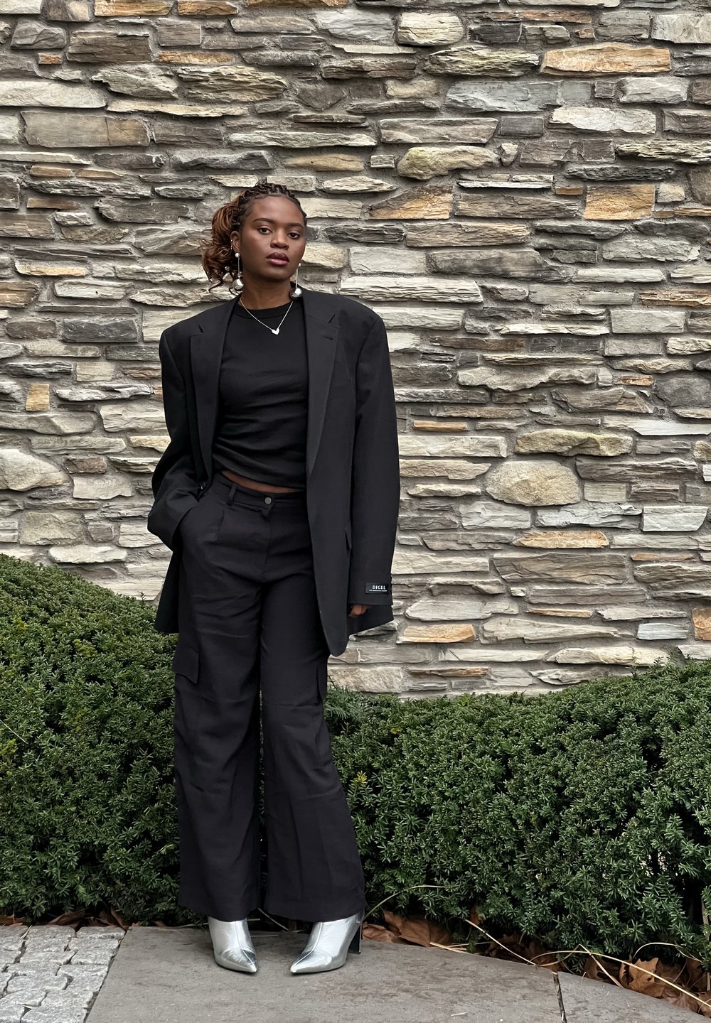 Zwarte oversized blazer over een zwarte crop top en wijde cargo broek. Zilveren high-heeled laarzen maken de look compleet. Textuur van een stenen achtergrond.