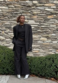 Zwarte oversized blazer over een zwarte crop top en wijde cargo broek. Zilveren high-heeled laarzen maken de look compleet. Textuur van een stenen achtergrond.