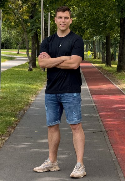 Camisa negra de manga corta, pantalones cortos de denim desgastados y zapatillas beige. De pie en un camino sombreado con hierba a un lado y una pista roja al otro.