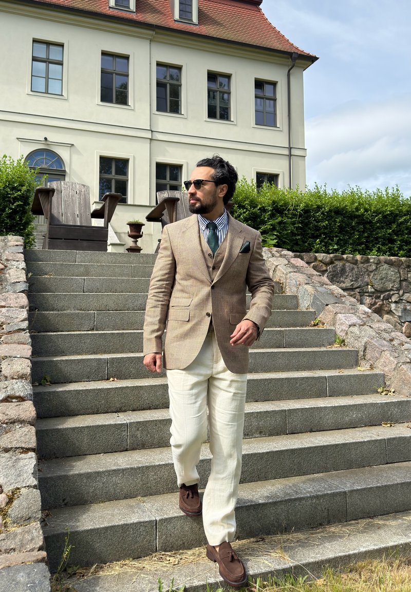 Man in beige blazer, white trousers, and sunglasses walking down stone steps outside a large cream-coloured house with red roof.