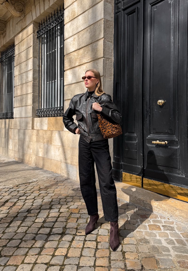 Fekete bőrdzsekit, fekete nadrágot és barna csizmát viselő nő, aki egy leopárdmintás táskát tart, a kőépület melletti macskaköves utcán áll.