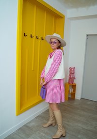 Roosa riie ruudulise mustriga pika varrukaga kleit, valge vest, beežid põlvekõrgused saapad ja lavendli värvi käekott kollase seina taustal.