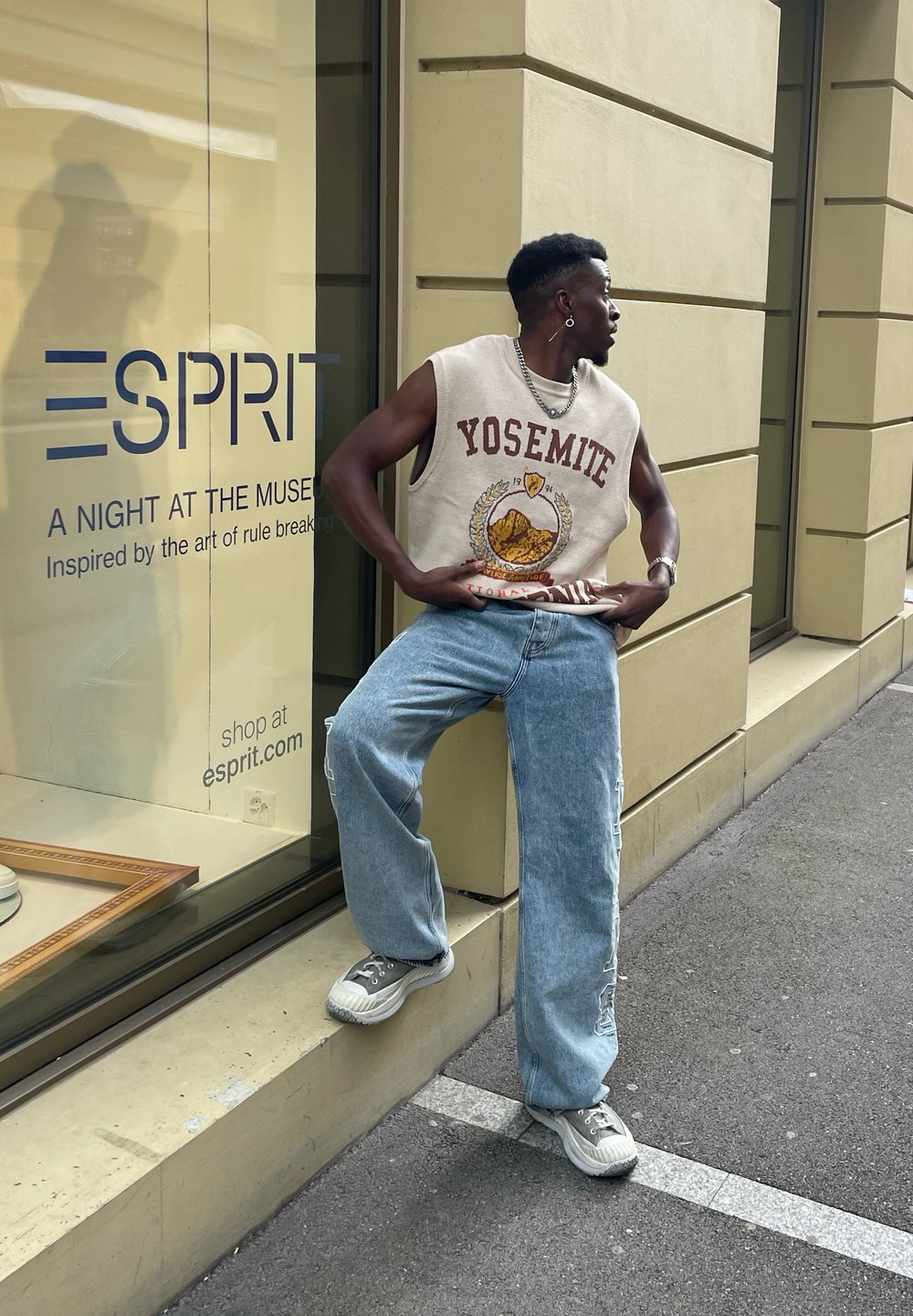 Szürke ujjatlan póló "Yosemite" grafikával, világoskék bő szárú farmernadrág és szürke sportcipő. A háttérben Esprit bolt kirakata látható.