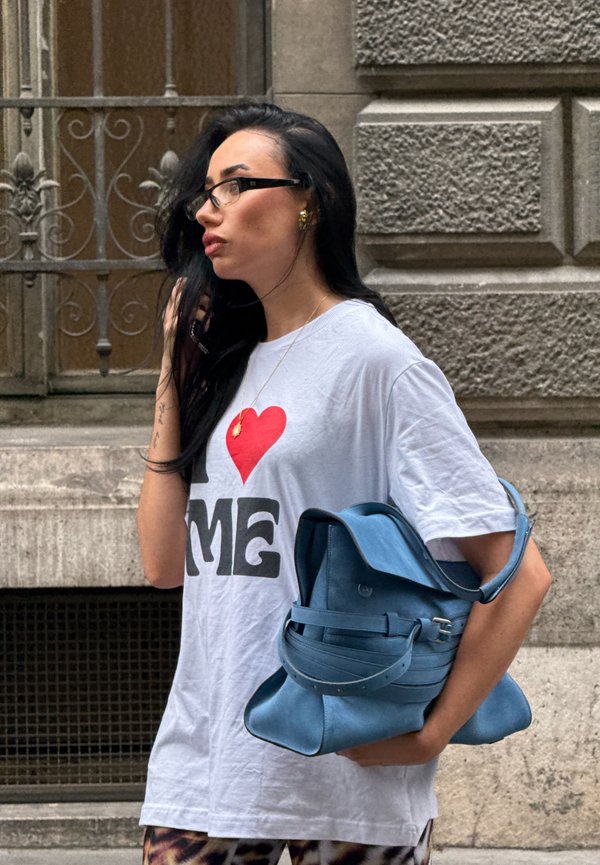 Femme aux longs cheveux foncés, portant des lunettes et un t-shirt blanc avec le texte "I love me", tenant un grand sac à main bleu dans une rue de la ville.