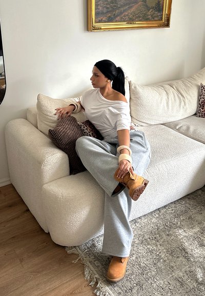 Mujer con coleta negra que lleva un top blanco de hombros descubiertos y pantalones grises, sentada en un sofá crema, sosteniendo una bota marrón claro, junto a cojines y alfombra con estampado.