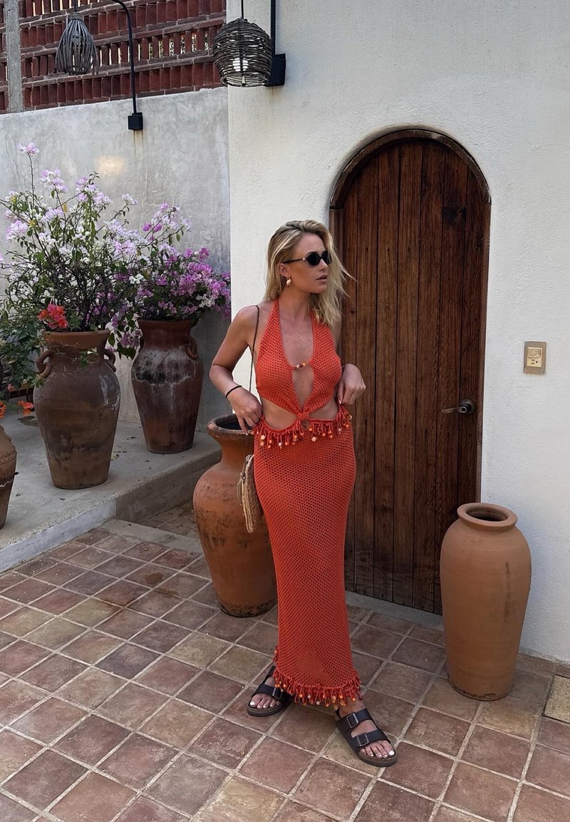 Woman wearing an orange crochet dress and black sandals stands near wooden arched door surrounded by large clay pots and flowering plants.
