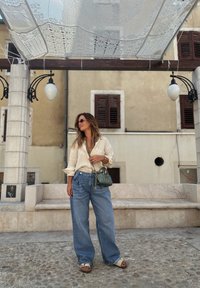 Camicia a maniche lunghe beige chiaro, pantaloni larghi in denim blu, borsa verde con texture, abbinata a sandali chiari, in piedi su pavimentazione in pietra.