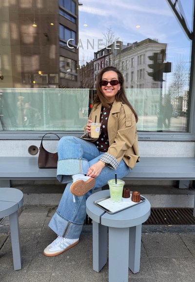 Mujer con gafas de sol y chaqueta beige sentada en un banco fuera de una cafetería, sosteniendo un café con hielo; bebida verde y pasteles en una mesa pequeña cerca.
