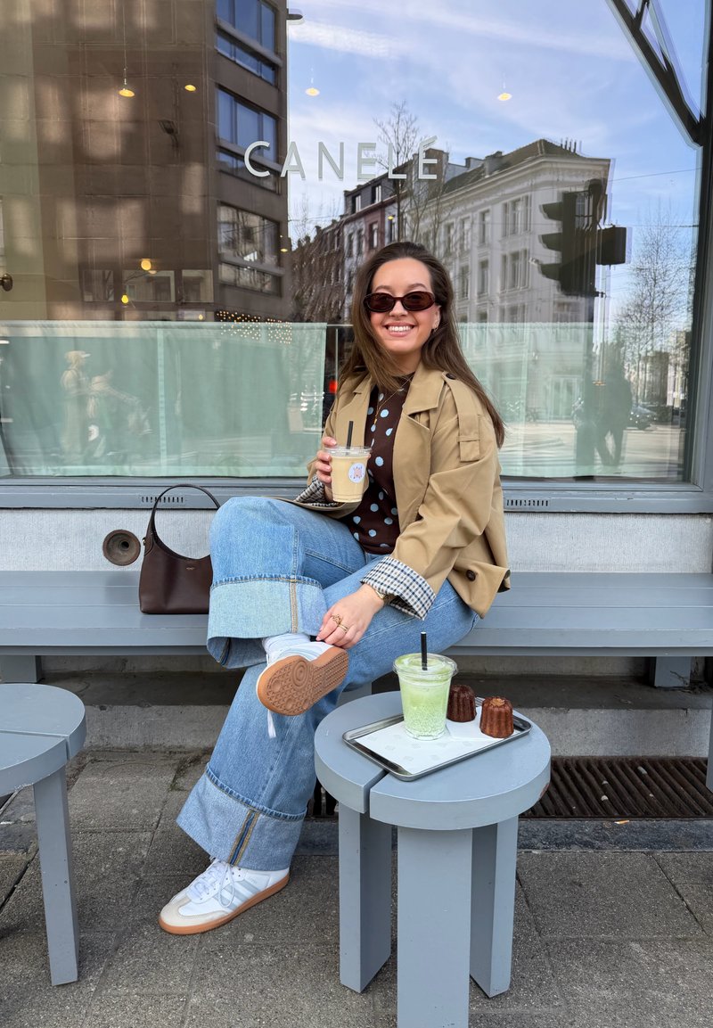 Femme portant des lunettes de soleil et une veste beige assise sur un banc devant un café, tenant un café glacé ; boisson verte et pâtisseries sur une petite table à proximité.