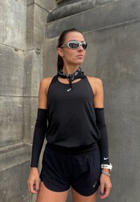 Débardeur de sport noir avec un design découpé, longues manches noires, shorts noirs et bandana noir. Lunettes de soleil argentées, montre-bracelet visible.